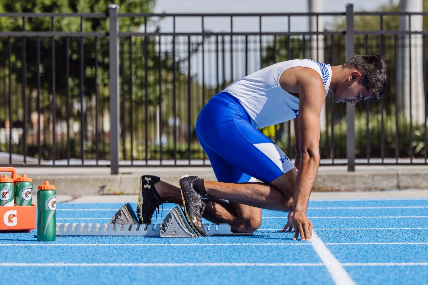 A track athlete in a white and blue uniform crouched in starting blocks on a blue running track with Gatorade bottles nearby.