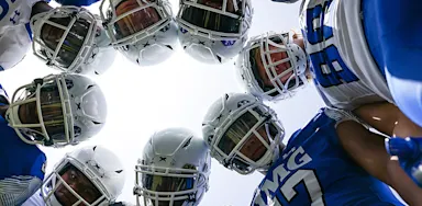 A group of football players in helmets and IMG uniforms huddle in a circle, looking down toward the camera during a team moment on the field.