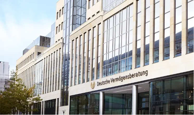 Modern office building with large glass windows and beige stone facade. Sign reads "Deutsche Vermögensberatung".