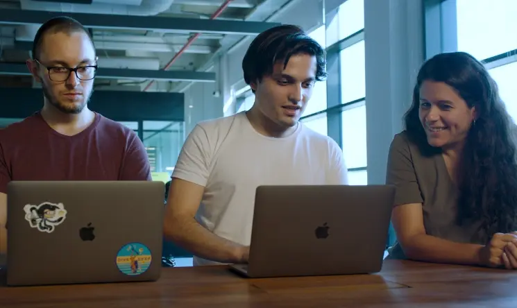 Three people seated at a table with laptops; one has a sticker-covered laptop, another in a white shirt is showing something on their screen to the third person.