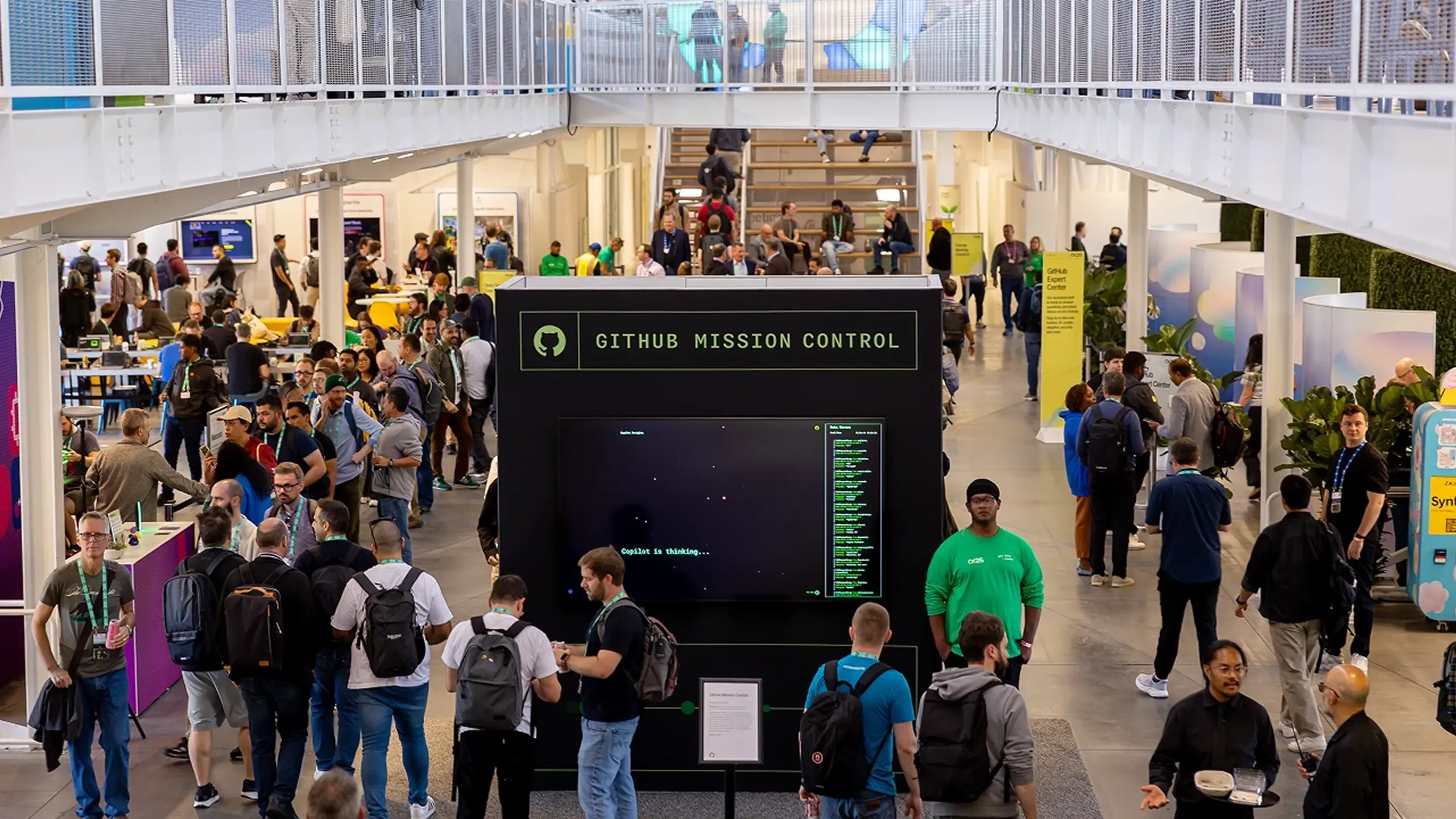 Attendees explore a busy exhibition hall around a “GitHub Mission Control” display, beneath colorful geometric graphics.