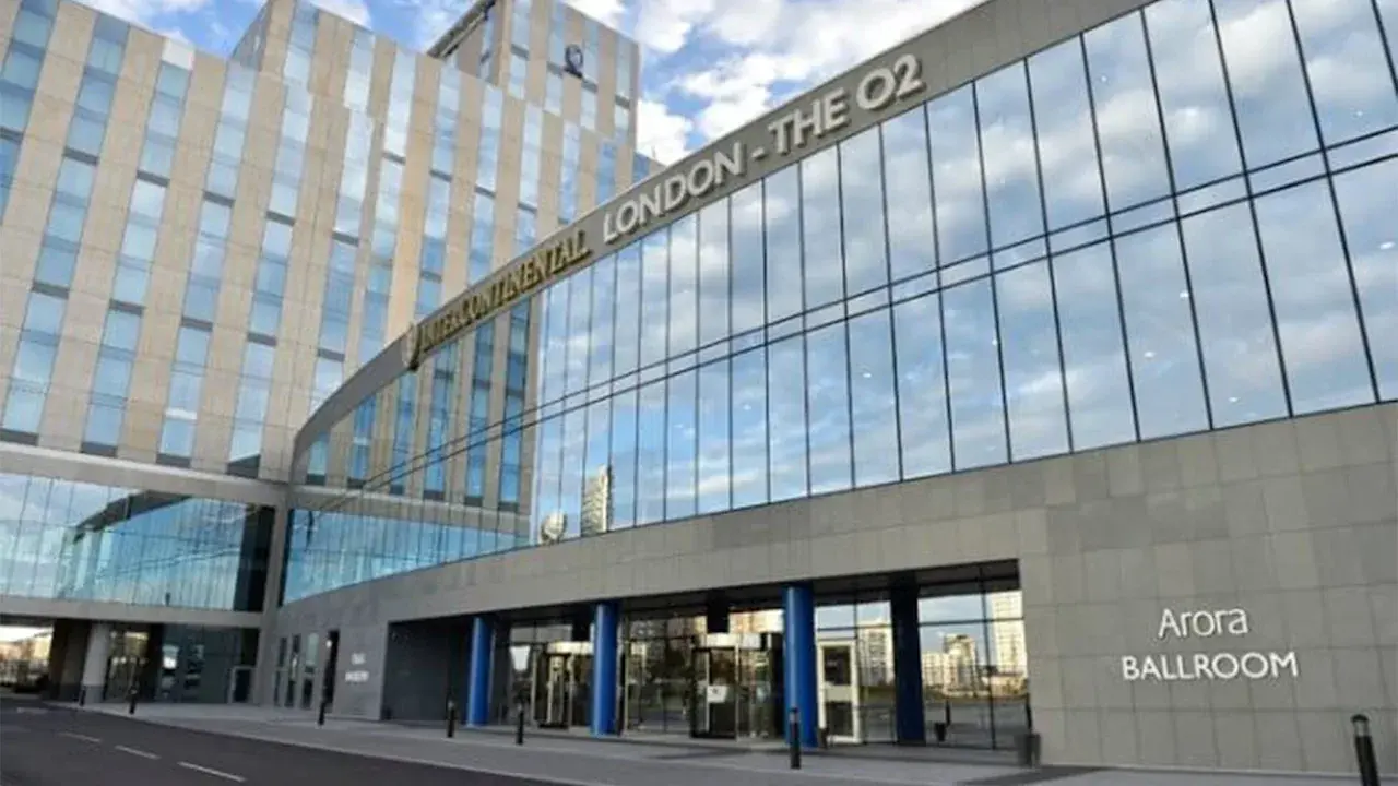 Exterior view of the InterContinental London - The O2 hotel, featuring a modern design with large glass windows reflecting the sky and nearby buildings. The hotel name is prominently displayed at the top, and a sign for 'Arora BALLROOM' is visible near the entrance.