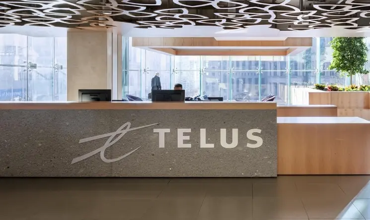 Modern office reception with gray TELUS desk, wood accents, decorative ceiling, and glass walls revealing an urban view.