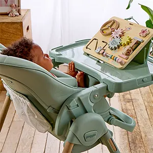 Toddler sitting in the pistachio green Puggle Feed & Play highchair, reaching towards the wooden toy stand on the tray.