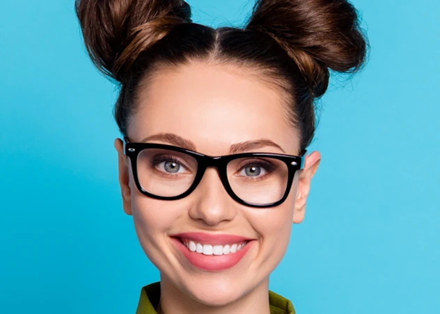 A woman withy fair skin, glasses and two dark brown space buns smiles forward in front of a light blue background.