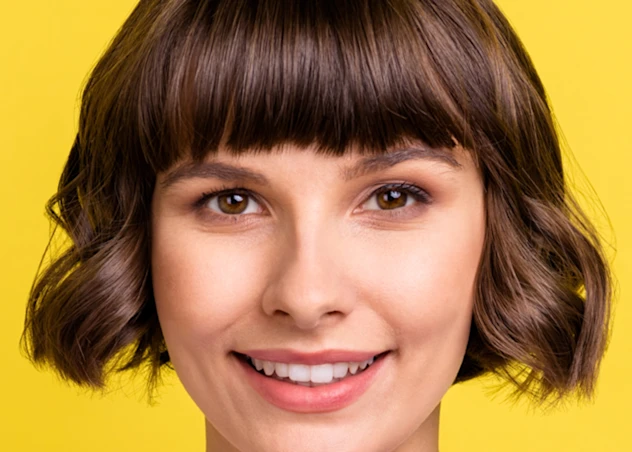 a woman with fair skin and a short brown bob hairstyle with bangs looks forward and smiles in front of a yellow backgound