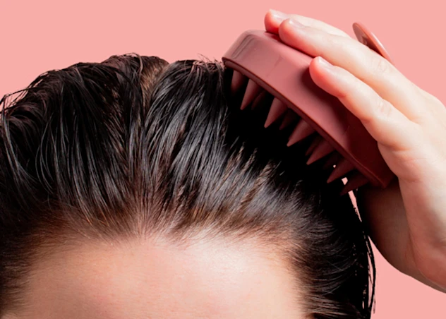 Woman with long dark hair and fair skin uses a brush to clean and exfoliate her scalp in front of a light pink background.