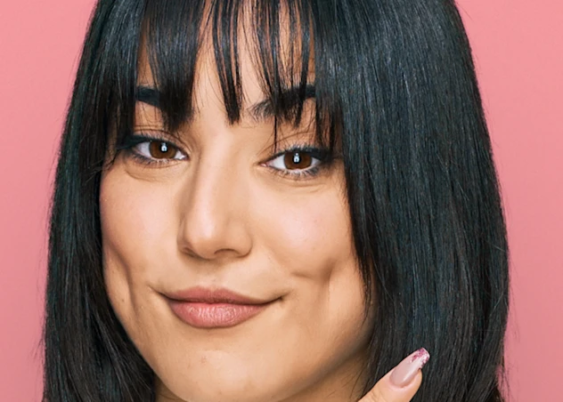 A woman with tan skin and medium length black hair and bangs grins facing forward in front of a pink background.