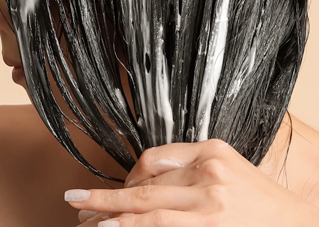 A tan-skinned woman applies hair masks to her black hair in front of a beige background.