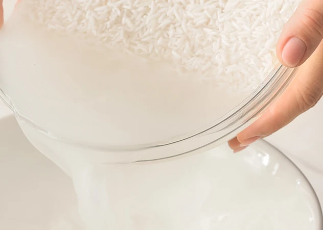 Fair-skinned hands pour rice water into a bowl from a bowl with rice in front of a white background.
