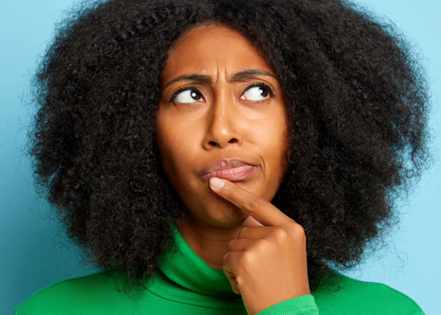 Dark skinned woman with natural 4c hair touches her face with her pointer finger giving an inquisitive look in front of a blue background