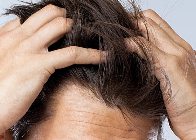 A fair-skinned man grabs his dark brown wavy hair in front of a grey background.