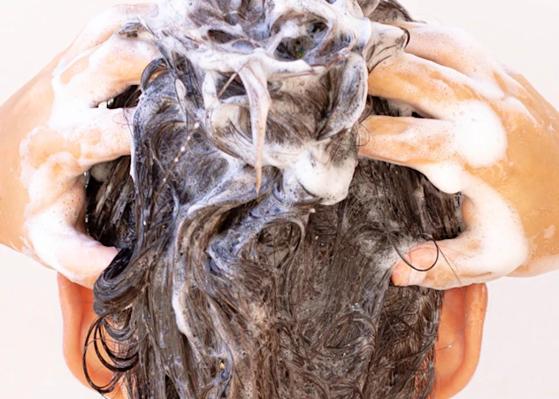 A person with tan skin washes their brown hair and scalp with foam in front of a white background.