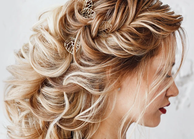 A woman with fair skin, wavy blonde hair in a prom hairstyle with leaf shaped clips looks down and right in front of a white background.