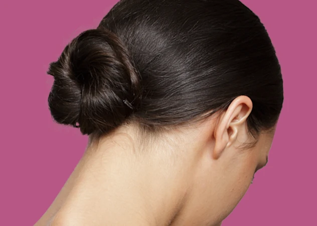 A woman with fair skin and dark brown hair in a bun looks down in front of a magenta background.