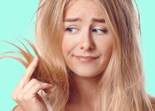 a fair skinned woman with damaged bleached hair holds it up with her fingers at looks at it with concern.
