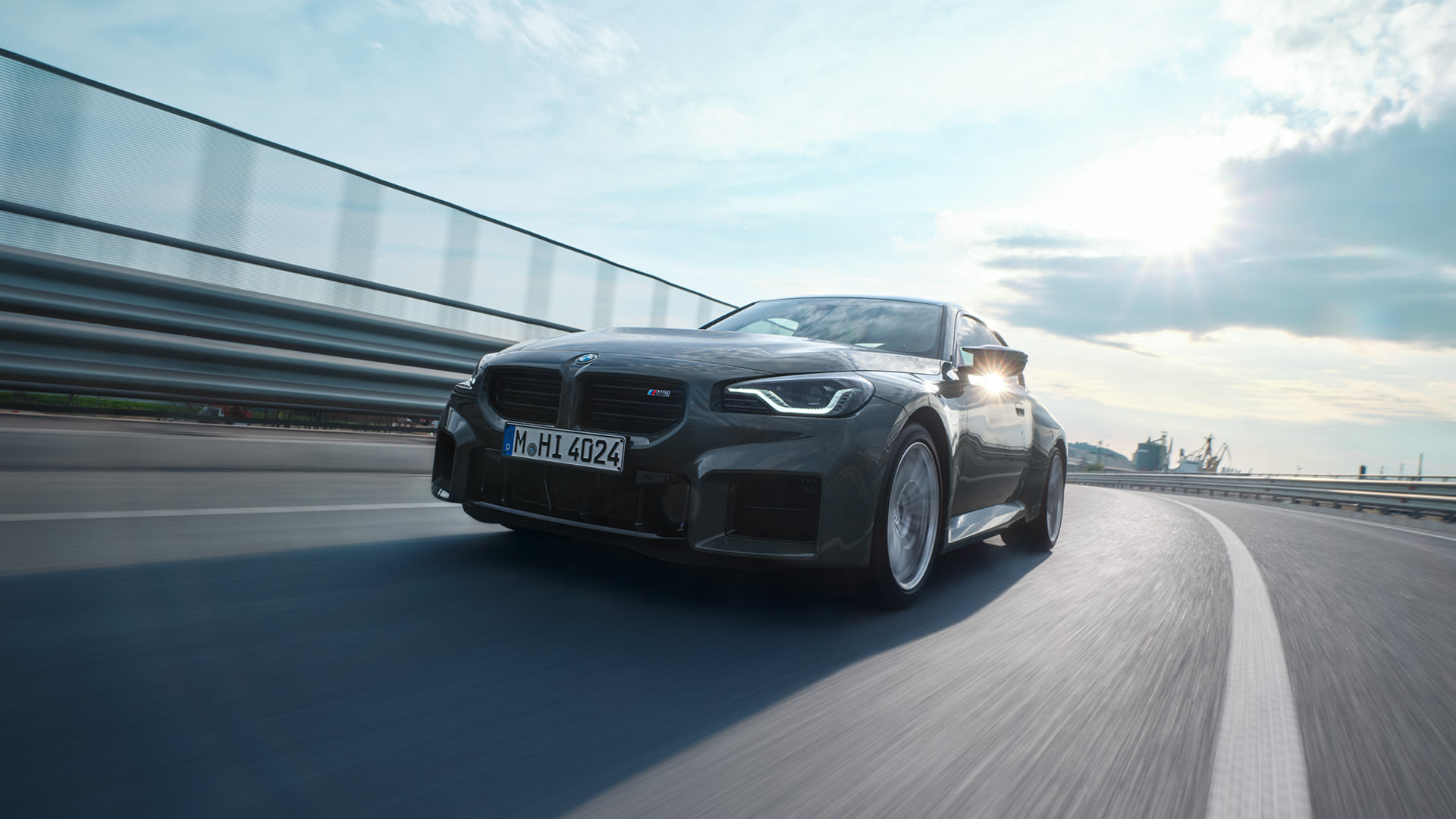 A black BMW car driving with a metal guardrail and a cloudy sky in the background.