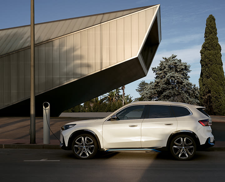 A white electric SUV is parked and charging at a public charging station in front of a modern, angular building with trees in the background.