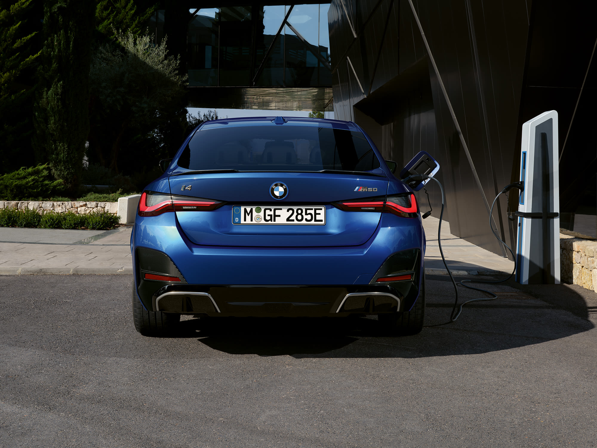 A blue BMW i4 electric car charging at a station in a modern urban setting with greenery and contemporary architecture in the background.