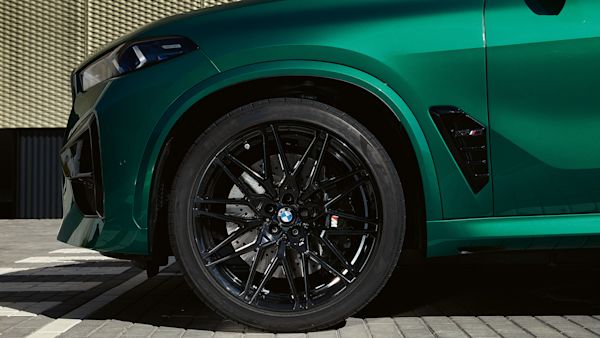 Close-up of the front side of a green BMW car with black alloy wheels, parked on a paved surface in front of a textured wall.