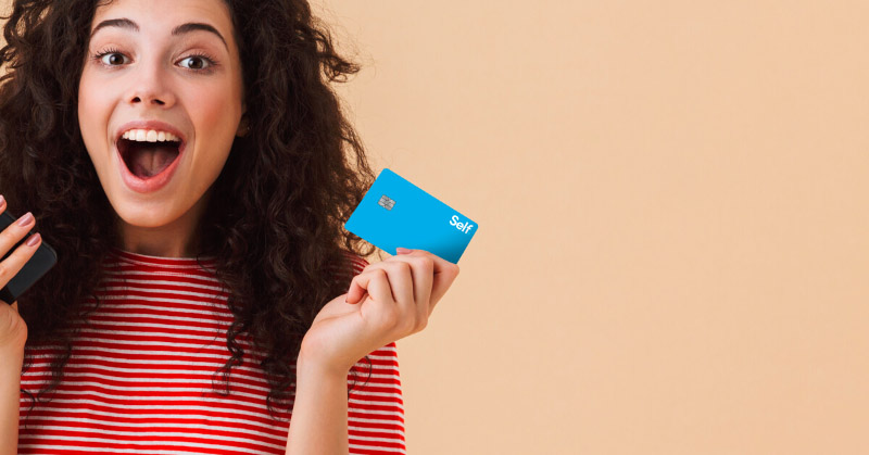 A young woman with curly hair and an excited expression shows off the bright blue Self Visa Credit Card. 