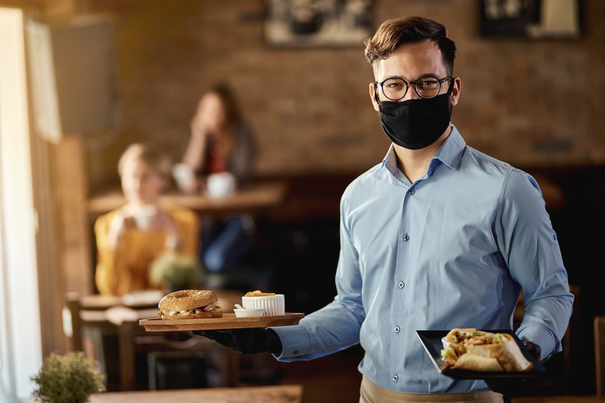 Young happy waiter wearing protective face mask while serving food