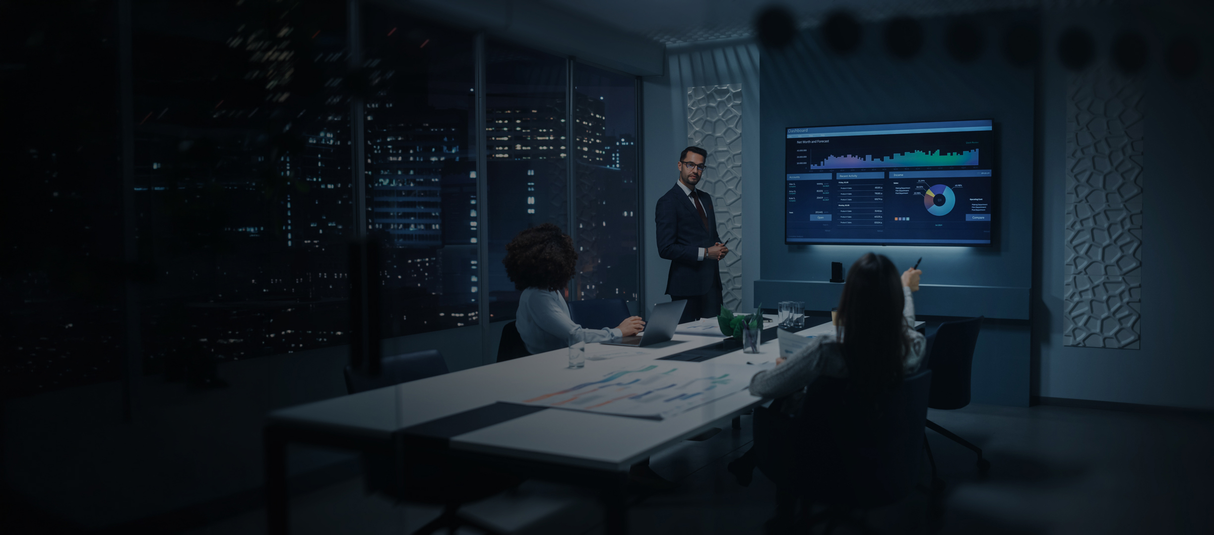 A professional man presents data on a large screen to two colleagues in a modern office and city lights can be seen through the window.