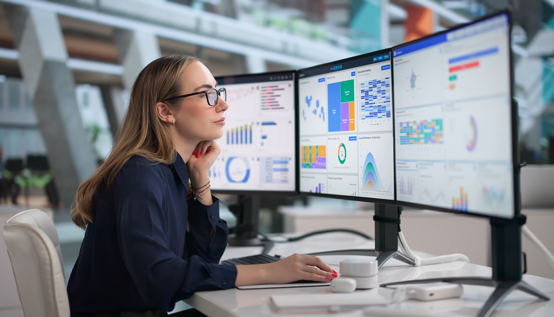 A woman with glasses looks at three monitors displaying data dashboards.