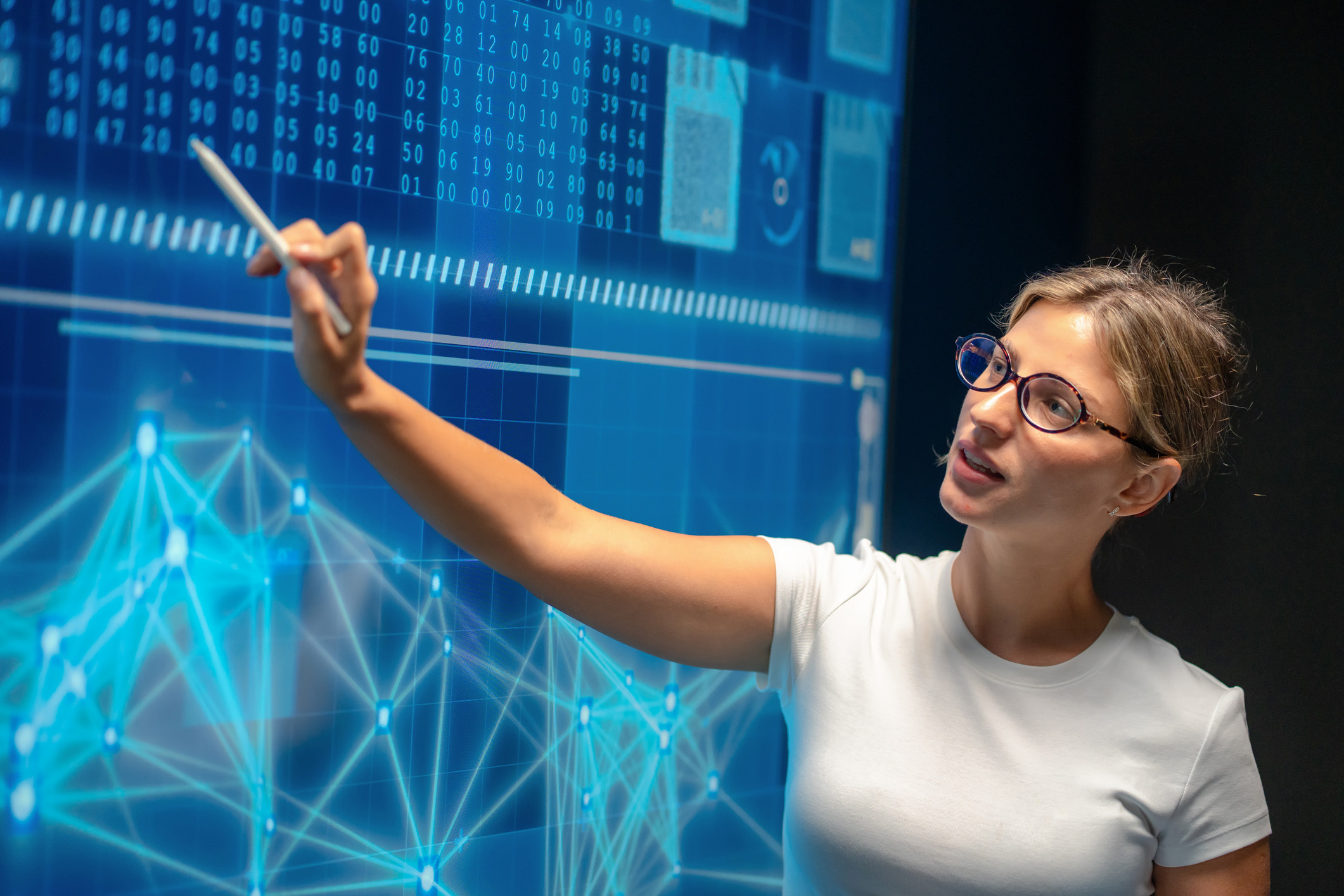 A woman wearing glasses holds a stylus and points towards a large, glowing blue digital interface in a dark room. The screen displays complex network graphs, charts, and lines of code that she appears to be analyzing.