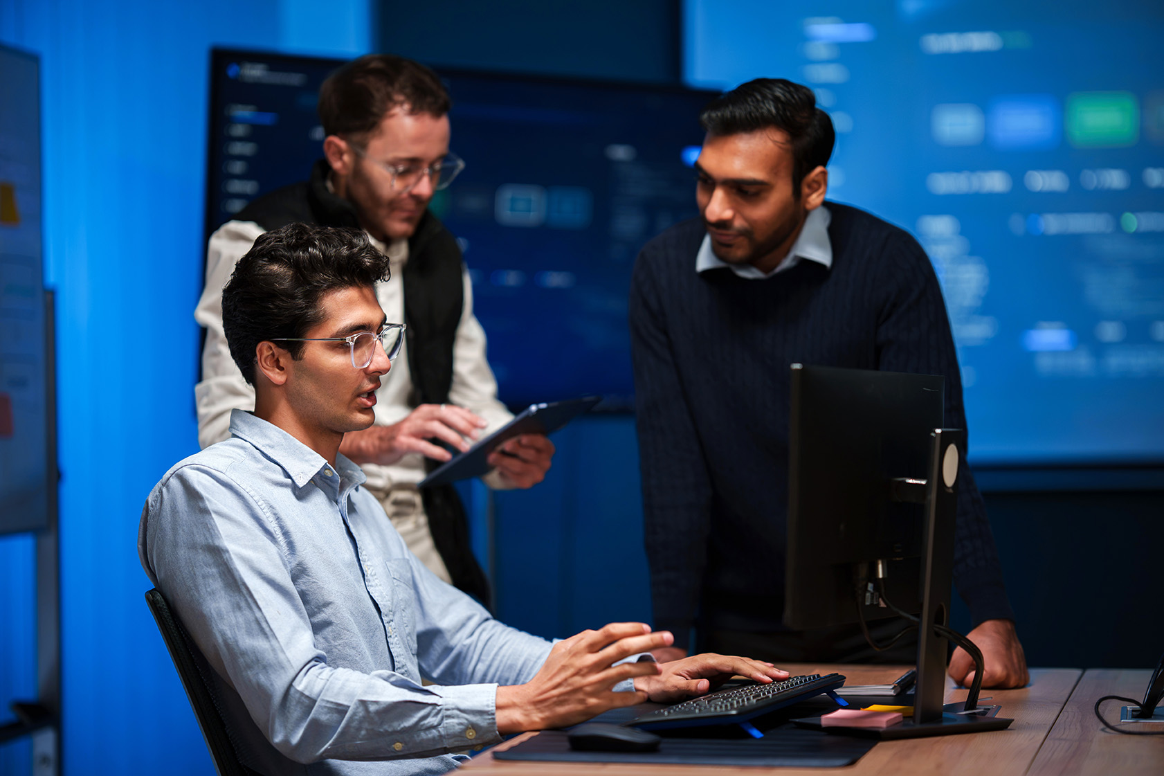 One man sits typing at a computer, while two other men stand behind him, all looking intently at the monitor and discussing workflows.