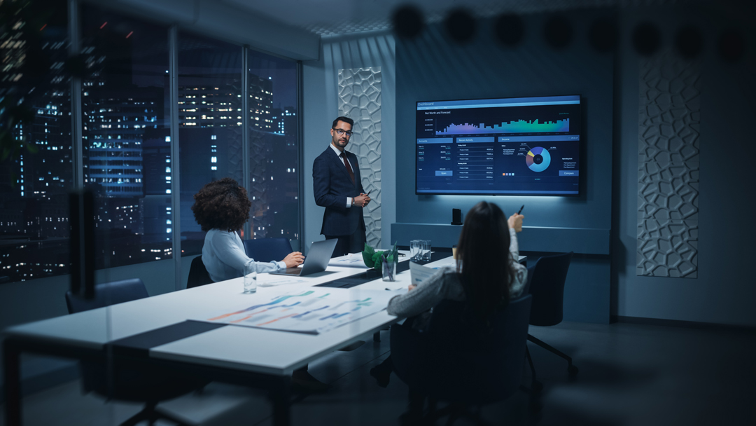 A professional man presents data on a large screen to two colleagues in a modern office and city lights can be seen through the window.