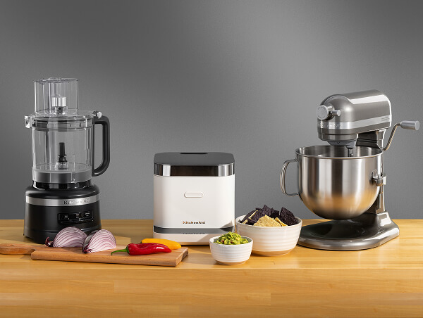 A black food processor, a white rice cooker and a silver stand mixer on a wooden counter surrounded by different ingredients in bowls and on a cutting board