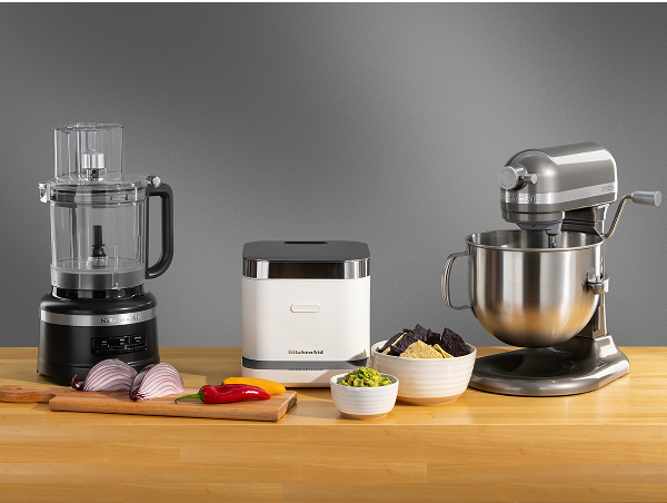 A black food processor, a white rice cooker and a silver stand mixer on a wooden counter surrounded by different ingredients in bowls and on a cutting board