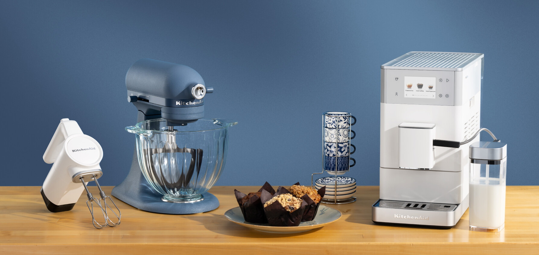 A white hand mixer, a Luminaire stand mixer and a white espresso machine on a wooden counter with muffins and mugs