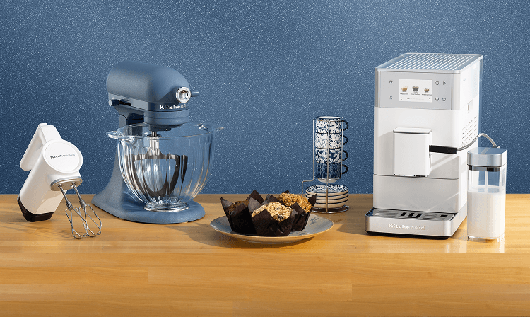 A white hand mixer, a Luminaire stand mixer and a white espresso machine on a wooden counter with muffins and mugs