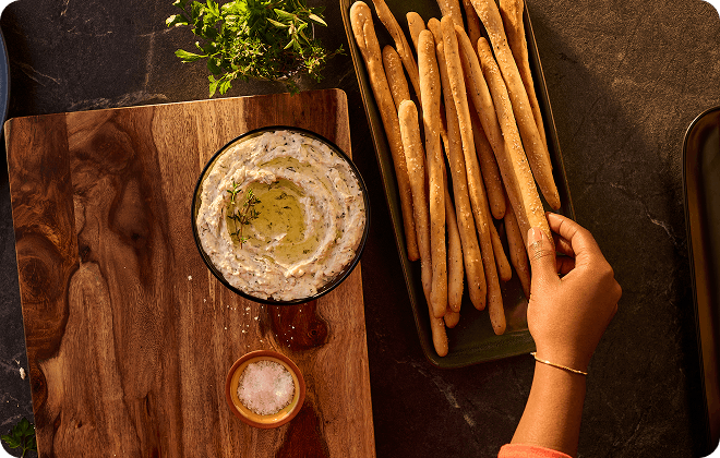 A person picking up a breadstick next to whipped feta dip.