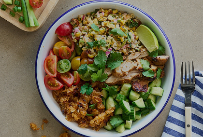 Elote Crispy Rice Salad topped with grilled corn, chicken, cherry tomatoes, cucumber, crispy rice, cilantro, and a lime wedge