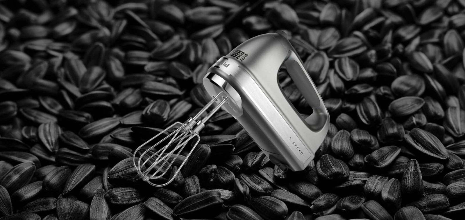 KitchenAid® hand mixer with a whisk attachment, set against a background of sunflower seeds