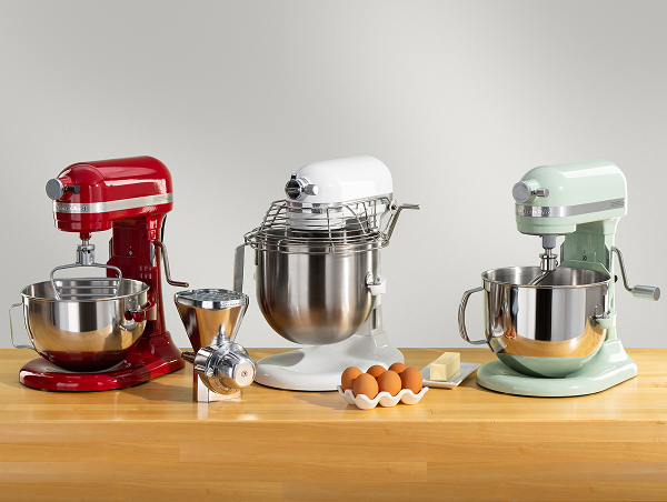 A trio of bowl-lift stand mixers in red, white and blue surrounded by different attachments and eggs on a wooden counter.