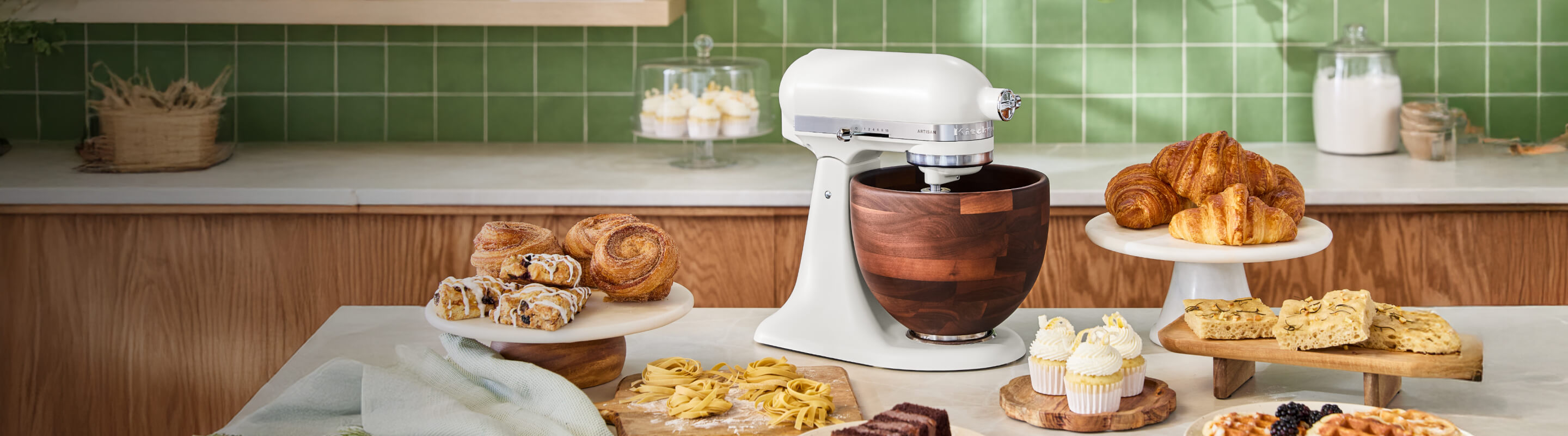 A Porcelain White KitchenAid® stand mixer with a wood bowl on the counter, near a slew of pastries.