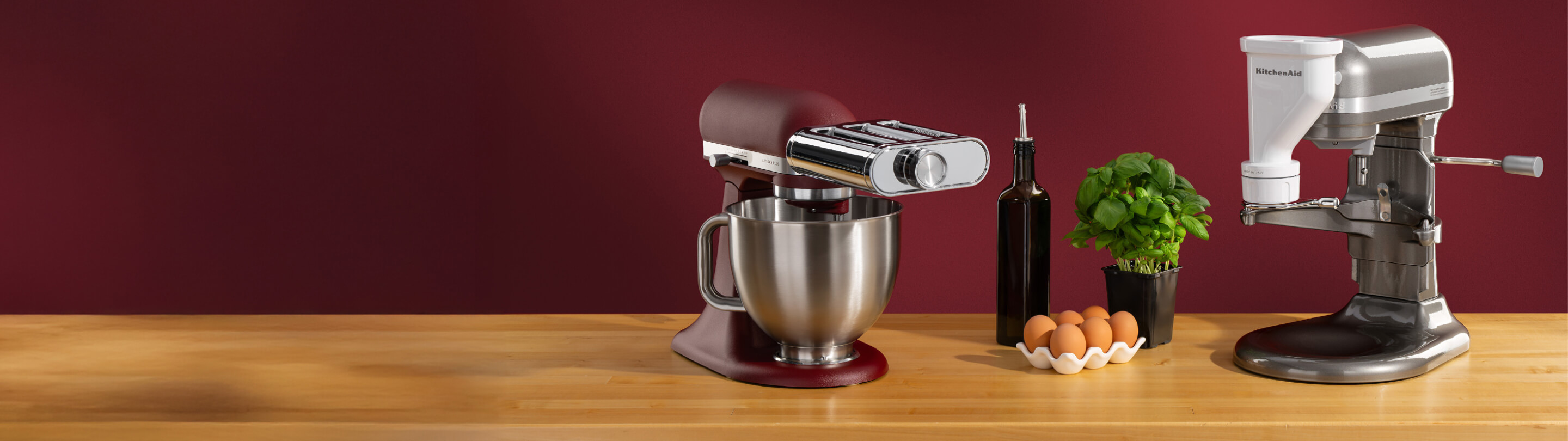 A red stand mixer with a pasta attachment and a silver stand mixer with a meat grinding attachment on a wooden counter with eggs, olive oil and basil