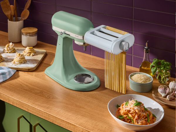 Green KitchenAid® stand mixer with a pasta roller attachment producing fettuccine, beside a bowl of pasta with tomato sauce