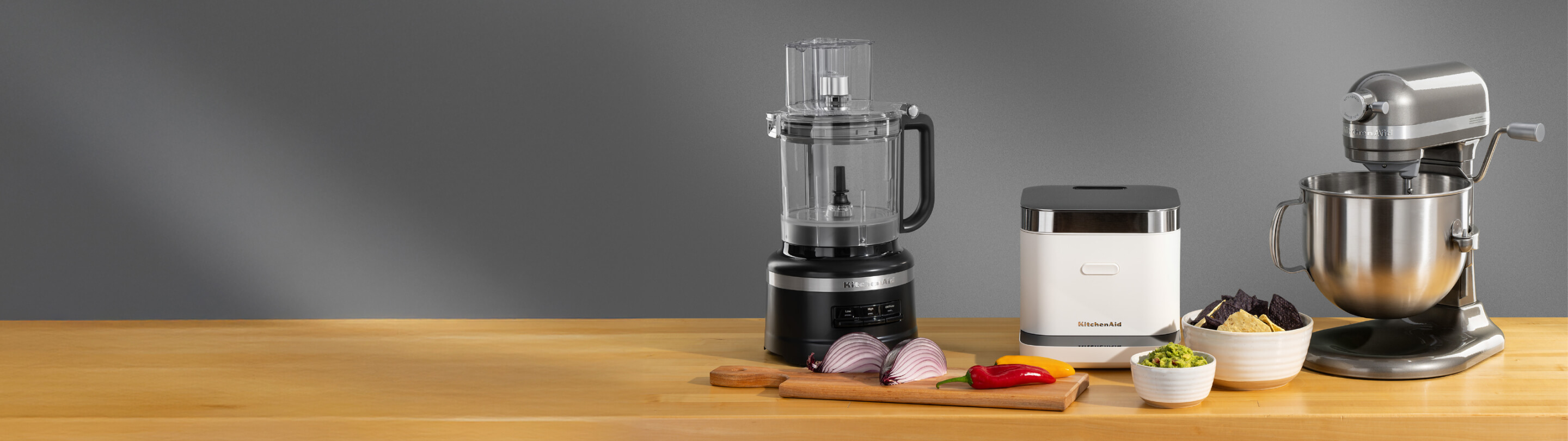 A black food processor, a white rice cooker and a silver stand mixer on a wooden counter surrounded by different ingredients in bowls and on a cutting board