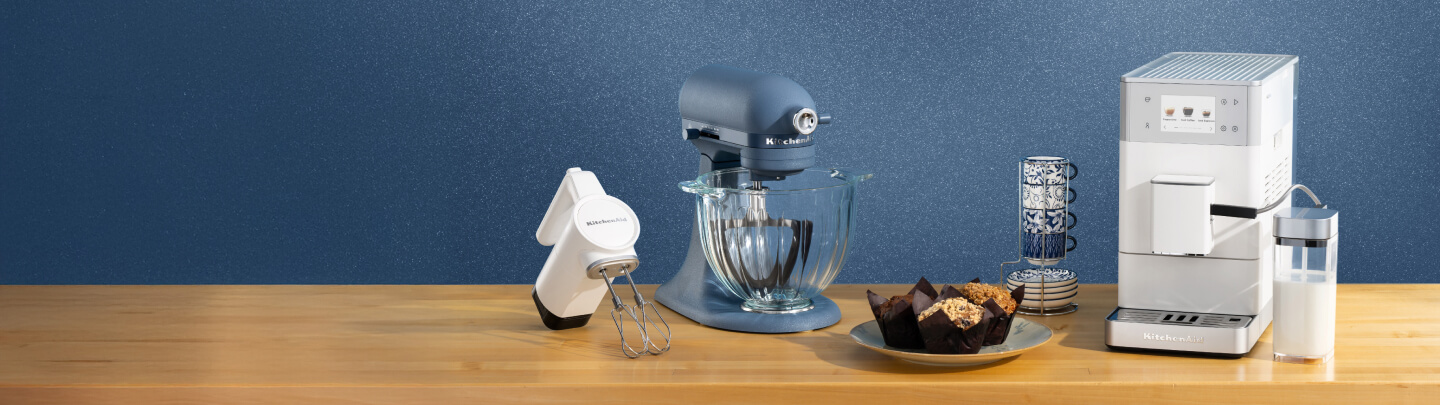 A white hand mixer, a blue stand mixer and a white espresso maker on a wooden counter with mugs and muffins