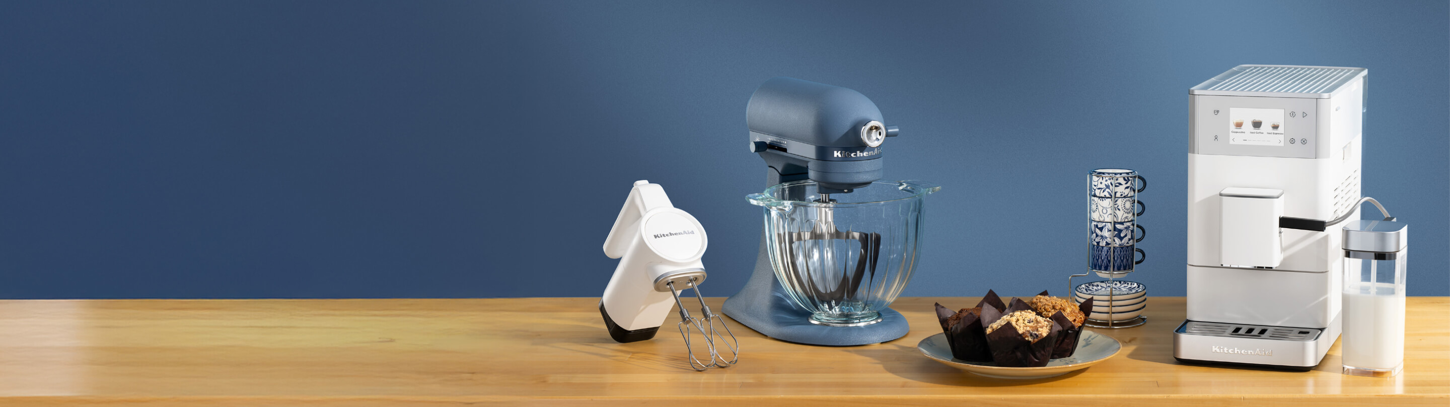 A white hand mixer, a blue stand mixer and a white espresso maker on a wooden counter with mugs and muffins