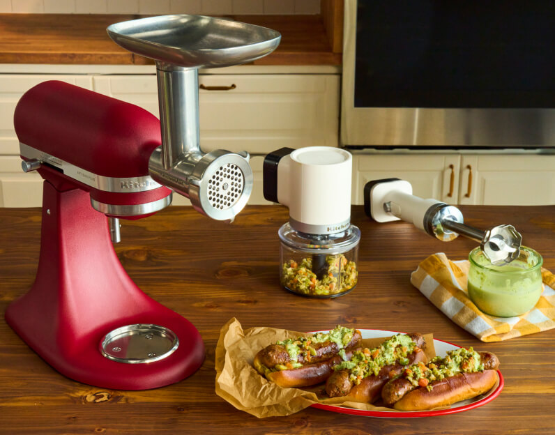 A KitchenAid® stand mixer with the Food Grinder attachment next to a plate of sausage.