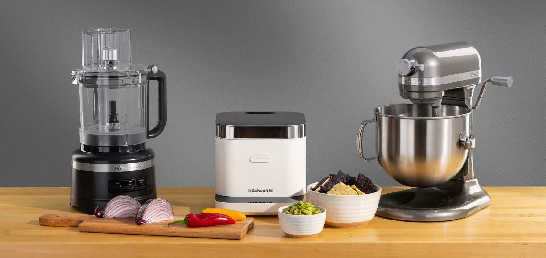 A black food processor, a white rice and grain cooker and a gray stand mixer on a wooden counter surrounded by fresh ingredients