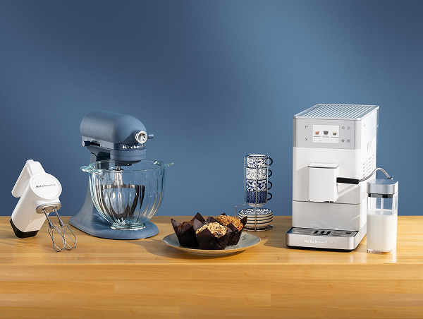 A white hand mixer, a blue stand mixer and a white espresso maker on a wooden counter with mugs and muffins
