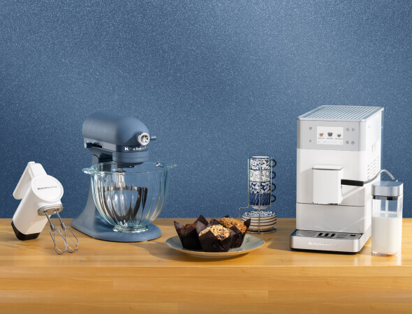 A white hand mixer, a blue stand mixer and a white espresso maker on a wooden counter with mugs and muffins