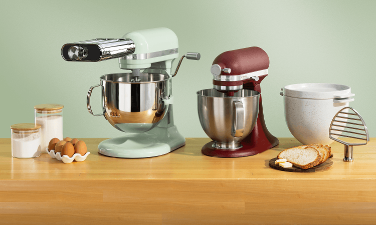 Green KitchenAid® stand mixer with a pasta roller attachment and burgundy stand mixer on a wood counter beside a ceramic bread bowl